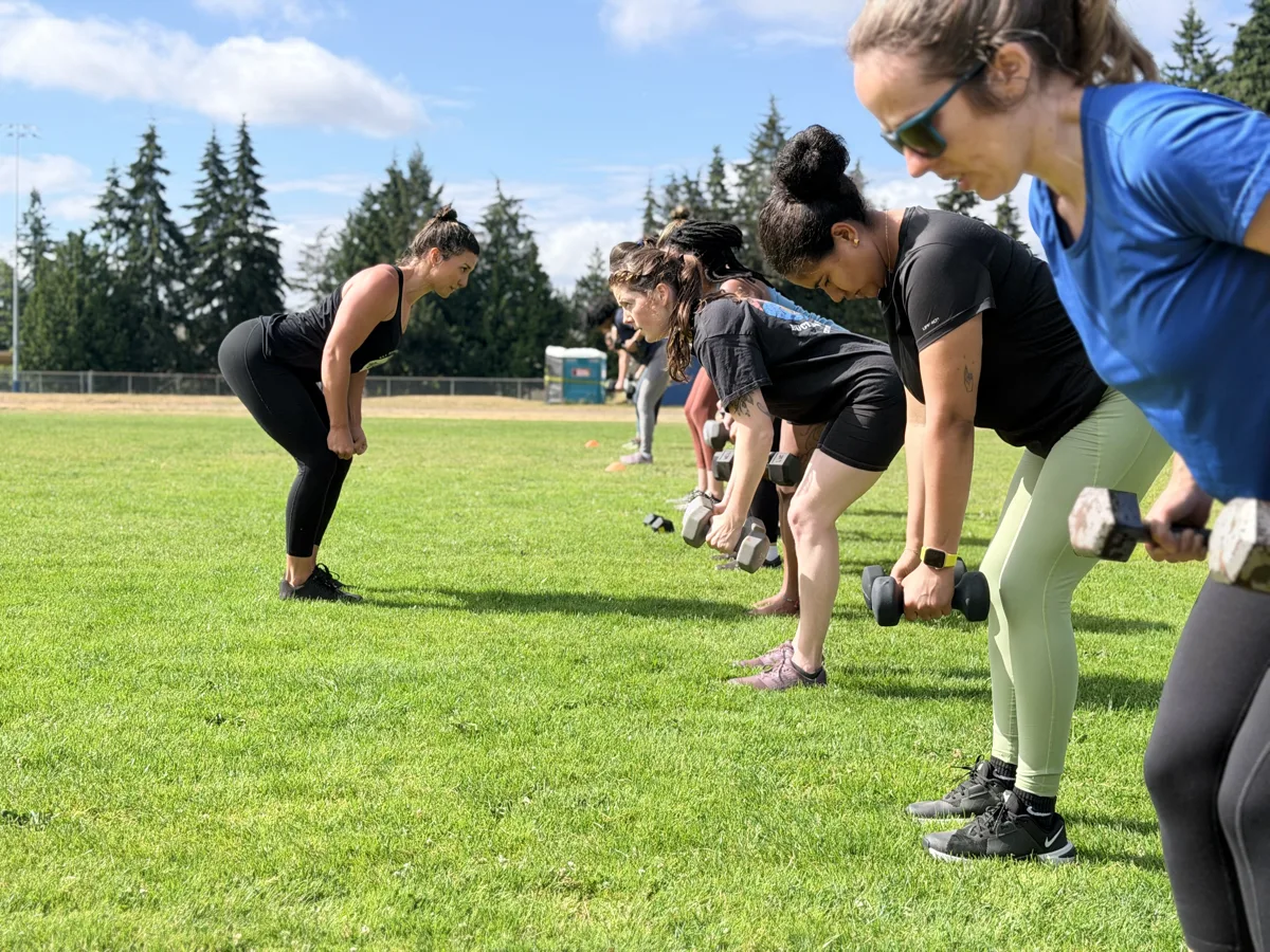 Coach Brittany demonstrates the hinge position while coaching a member through dumbbell rows at an outdoor class